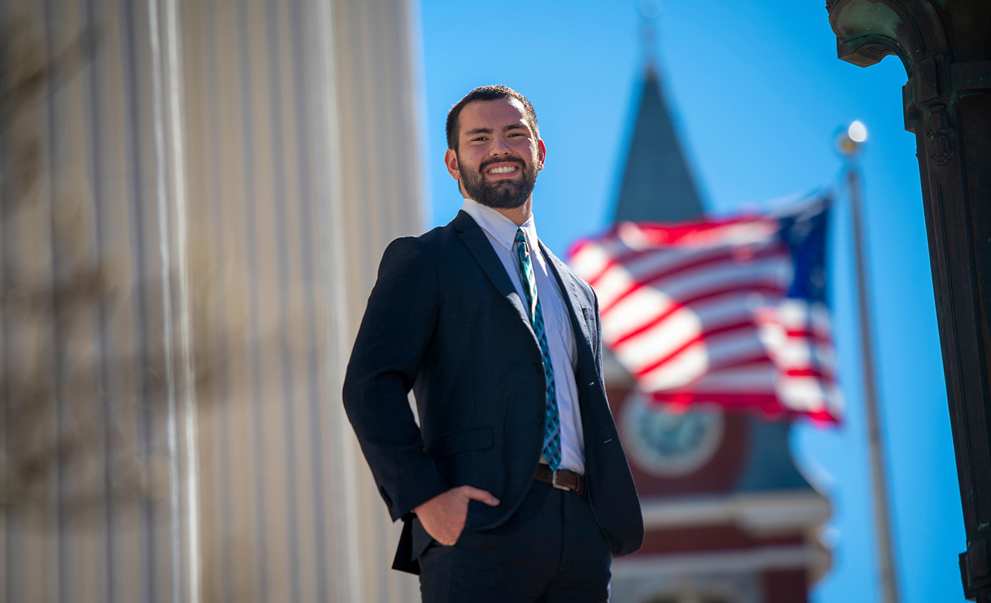 UNCW student standing at Wilmington City Hall with the American flag and spire of the New Hanover County Courthouse in the background.