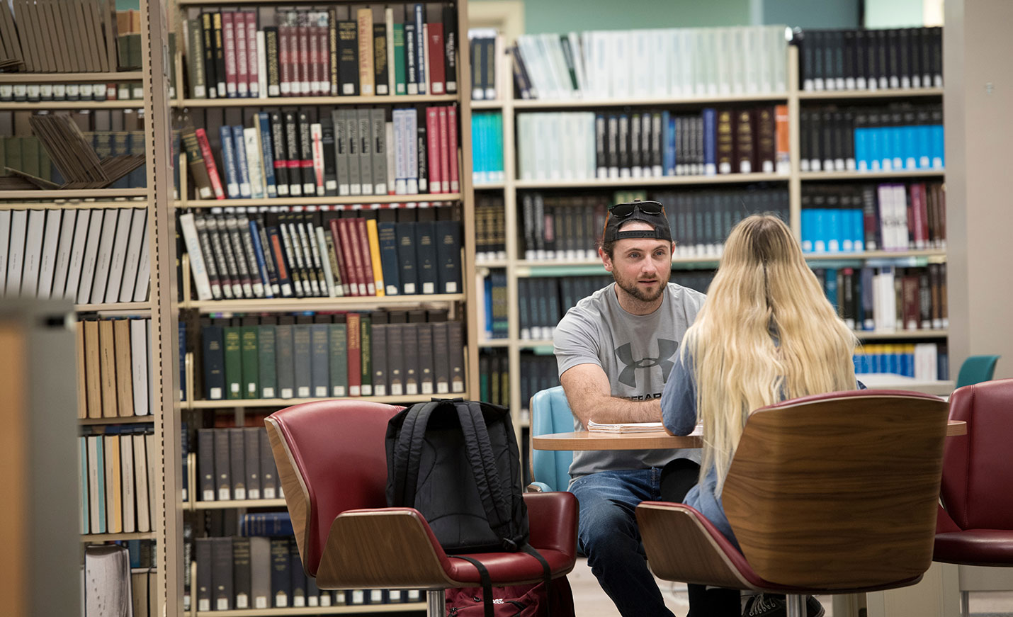 Two students talk while sitting across a table from each other in the library