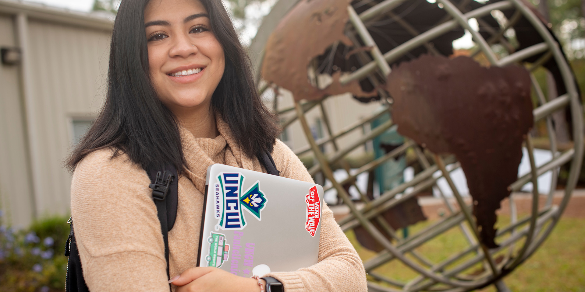 A student holding a laptop stands in front of a statue of a globe