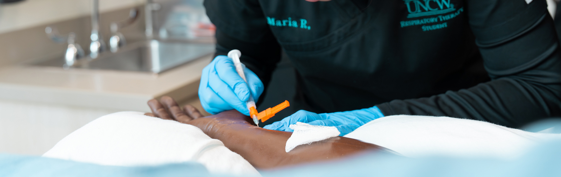 the gloved hands of a respiratory therapy student practices drawing blood from the radial artery of a manikin. 
